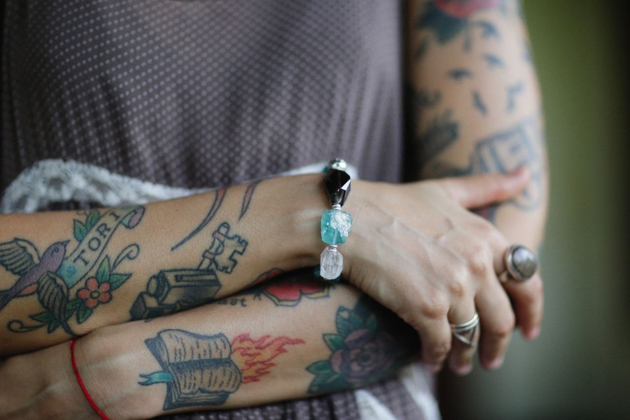 our-mission-01 Close-up view of tattooed arms with a bracelet and rings, showcasing unique body art and style.