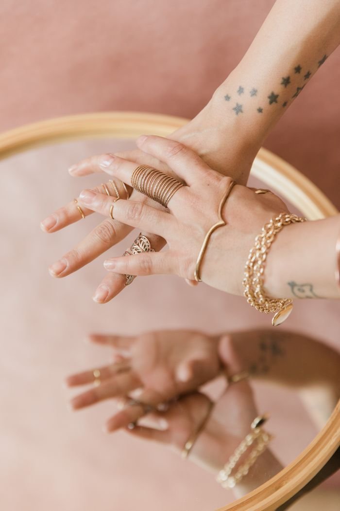 Close-up of tattooed hands with gold rings and bracelets on a reflective surface.